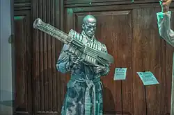 Statue de cire, argentée, représentant un homme chauve en peignoir tenant un fusil de science fiction.