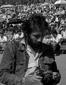 Photographie en noir et blanc, portrait en buste. Un homme aux cheveux mi-longs et avec une barbe regarde l'appareil photo qu'il tient dans ses mains.