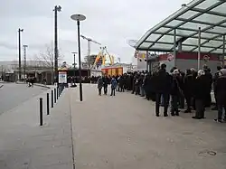 File d'attente pour l'accès aux navettes spéciales pour les journées portes ouvertes des travaux de la ligne B du métro. à gauche, les arrêts de bus. En arrière-plan, la fête foraine. À droite, l'auvent abritant des commerces.