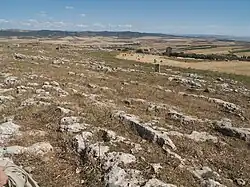 Vue d'alignements de pierre au premier plan avec à l'arrière-plan une ville en ruines et au fond un paysage vallonné