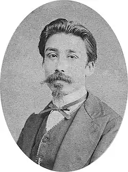 Photographie en noir et blanc. Portrait en buste d'un homme élégant, avec les cheveux gominés en arrière, une longue moustache pointue et un bouc pointu.
