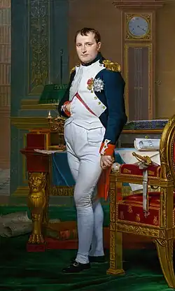 Photographie en couleurs du portrait en pied d'un homme portant une tenue militaire et ses décorations, glissant la main droite dans son gilet. En arrière-plan on distingue le mobilier d'une pièce peu spacieuse : un secrétaire et une horloge.
