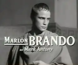 Photographie en noir et blanc d'un homme portant la toge, le regard pensif vers la bas de l'image. Des caractères de grande taille, en surimpression, indiquent que Marlon Brando joue le rôle de Marc Antoine.