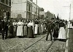 Dans une rue montante, un cortège de femmes nu-tête, en corsage et jupe longue, est surmonté d'un panneau « VIVE LE SUFFRAGE UNIVERSEL ».