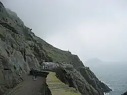 Photographie en couleurs d'un chemin taillé à flanc de falaise et délimité à droite par un parapet en pierre, un bâtiment installé à son extrémité.