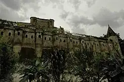 Photo couleur d'un rempart en brique rouge à trainées vertes de mousse. Le mur taluté en bas comporte des contreforts en demi-cylindre et le haut est doté d'une rangée de fenêtres sous le chemin de ronde. La tour à droite est polygonale surmontée d'un toit pointu, également en brique.