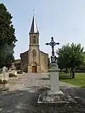 La tour clocher de l'église, au premier plan, une croix monumentale en fer forgé où est représenté au centre Jésus-Christ.