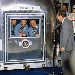 Photographie en couleur du président Nixon se tenant à côté d'un compartiment de quarantaine avec les astronautes visibles par la partie vitrée.