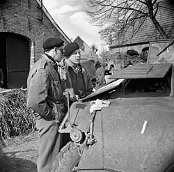 Photo noir et blanc montrant deux hommes en uniforme en train de discuter entre des maisons, près d'un véhicule blindé.