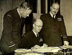 Trois hommes en tenue militaire, deux debout et un assis, regardant des papiers sur un bureau.