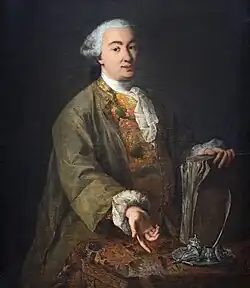 Portrait d'un homme, main gauche sur un livre posé à la verticale sur une table et tendant la main droite ouverte, paume vers nous qui le regardons.