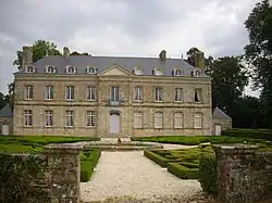 Photographie en couleur montrant la façade d'un château néo-classique, devant une cour avec une fontaine entourée de haies taillées.