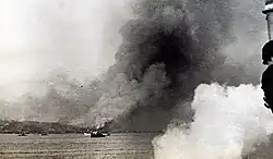 Photo noir et blanc d'un port avec beaucoup de navires et en fond, un immense panache de fumée.