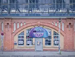 Image représentant l'entrée du club, niché dans les voutes en brique sous le S-Bahn berlinois. Le logo, formé d'une boule disco bleu-violette, surplombe la petite porte d'entrée en métal.