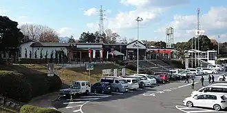 Blick auf einen niedrigen, modernen Gebäudekomplex, der leicht erhöht liegt und von Bäumen umstanden ist. Davor eine Betonfläche mit vielen parkenden Autos, in der Mitte der Fahrzeuge eine freie Fahrgasse mit weißen Pfeilen und einem Rollstuhl-Piktogramm.