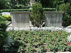 Frontale Farbfotografie von zwei einfachen Grabsteinen mit Nadelsträuchern neben ihnen. Auf dem linken Stein steht „Oona Chaplin 1925–1991“ und auf dem rechten Stein steht „Charles Chaplin 1889–1977“. Im Vordergrund sind in der Erde grüne und weiße Topfpflanzen. Im Hintergrund sind Gräber und eine Hecke.