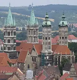 Von erhöhter Position aufgenommen. Der Dom hat vier Türme (grüne kupferne Spitzen), das Schiff des Doms hat ein Ziegeldach. Im Hintergrund sieht man die Hänge des Saaletals