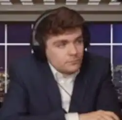 Ein Mann Anfang 20 mit braunen Haaren und braunen Augen. Er trägt ein weißes Hemd unter einem dunkelblauen Sakko und hat Kopfhörer auf.