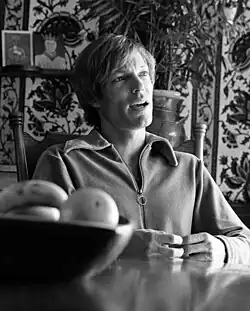 Schwarz-weiß Foto von Richard Chamberlain (1972) sitzt er an einem Tisch und trägt einen Pullover mit einem Reißverschluss. Vor ihm steht eine Schale, die mit mehreren Früchten, vermutlich Orangen, gefüllt ist. Der Hintergrund des Bildes zeigt eine Wand mit einem dekorativen Muster. Zudem sind einige Pflanzen sowie ein Regal sichtbar, auf dem ein gerahmtes Bild steht. Das gerahmte Bild zeigt eine Person mit einer Krone. Die gesamte Szene vermittelt eine ruhige, alltägliche Atmosphäre.