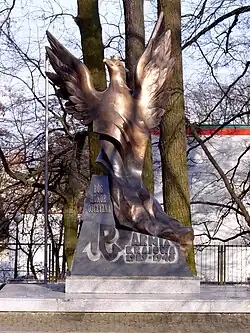 Zdjęcie ok. 4-metrowego pomnika, przedstawiającego orła z rozpostartymi skrzydłami. Na postumencie napisy: „Armia Krajowa 1939-1945”, „Bóg, Honor, Ojczyzna” oraz znak Polski Walczącej. Za pomnikiem kilka bezlistnych drzew, dalej za płotem znajduje się biały budynek. Zdjęcie wykonano w lekko słoneczny dzień.