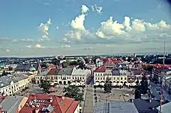 Na pierwszym planie Rynek Krosna z lotu ptaka. Zabytkowy plac z niewielką ilością drzew. Dookoła niewysokie kamienice.